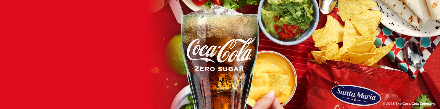 En glas med Coca-Cola-logga fyllt med Coca-Cola och is framför tacotillbehör som cheesedip, tortillachips, tortillabröd, guaccamole, lime, och koriander.