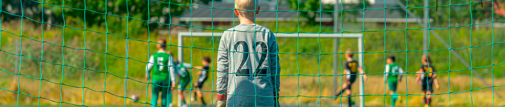 En pojke klädd i fotbollskläder står i ett fotbollsmål på en grön fotbollsplan. I bakgrunden skymtas andra barn som också spelar fotboll.