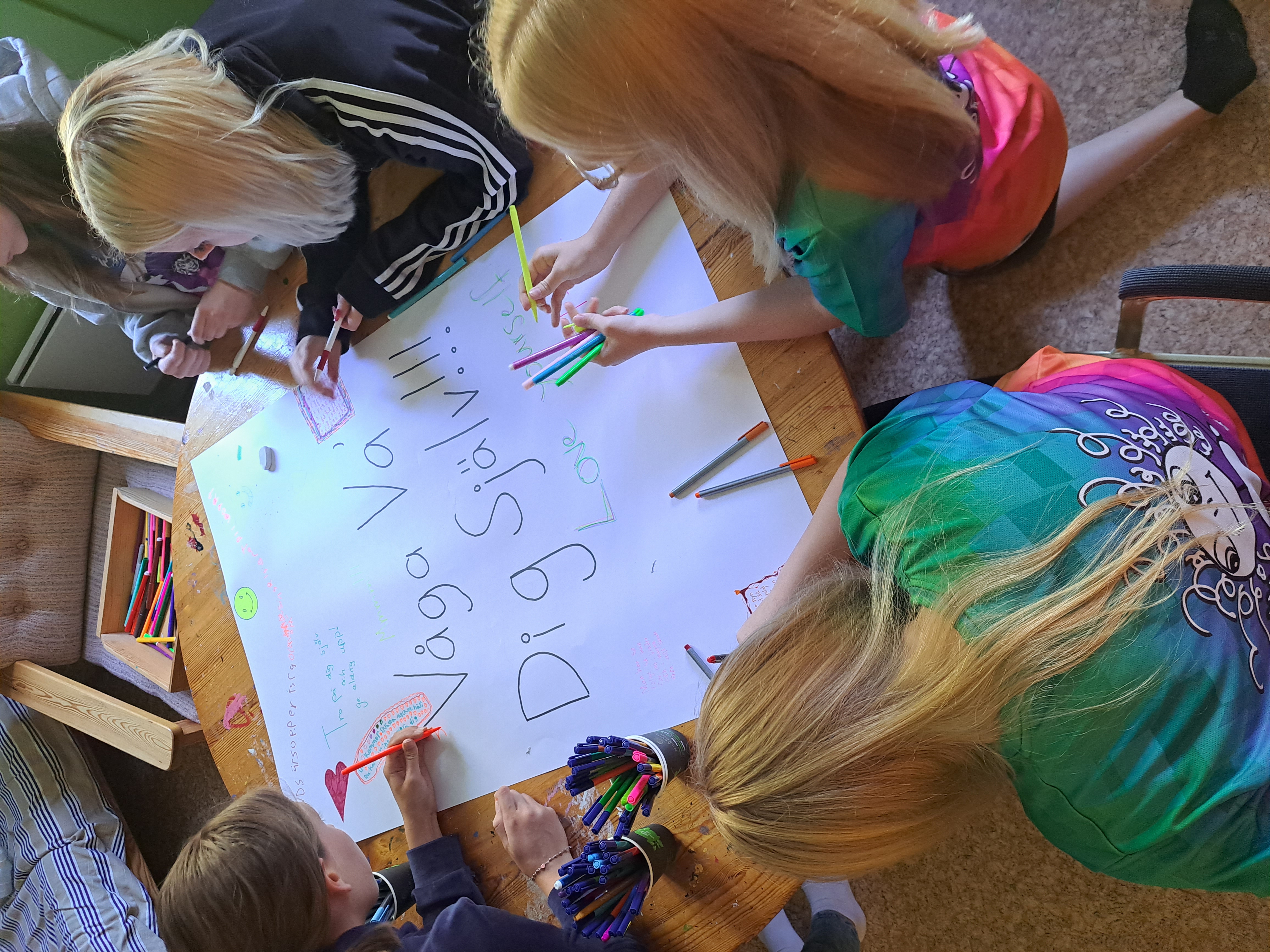 Flera barn sitter vid ett träbord och målar med färgglada pennor på ett stort vitt papper. 