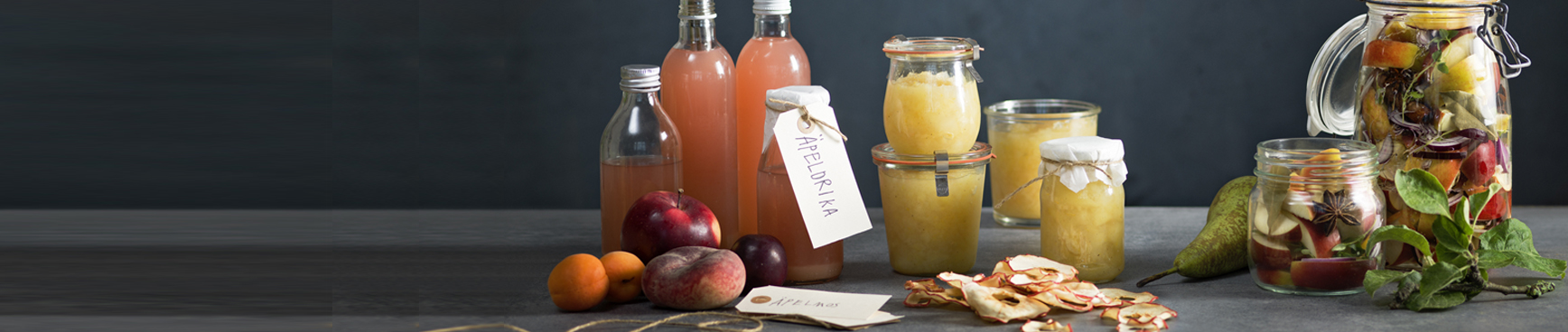 Glasflaskor med äppeldricka och glasburkar med äppelmos står bredvid en hög med röda äpplen på ett grått bord framför en mörk bakgrund.